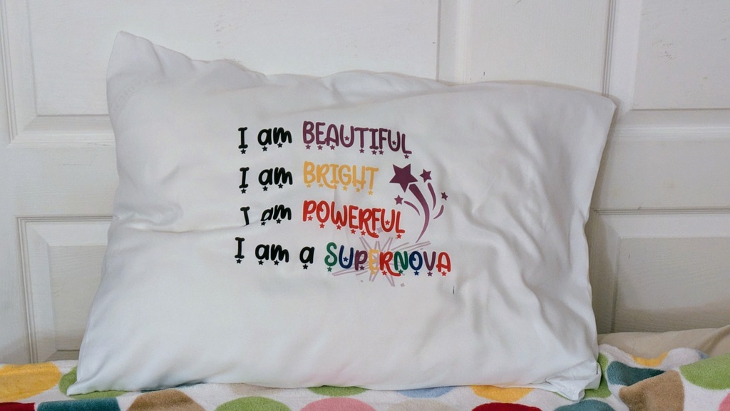 The finished pillowcase, with a pillow inside on a bed. The image reads:
I am BEAUTIFUL
I am BRIGHT
I am POWERFUL
I am a SUPERNOVA
with small stars under each letter. The words are colored in purple, yellow, and red, while supernova is has each letter in a different bright color.